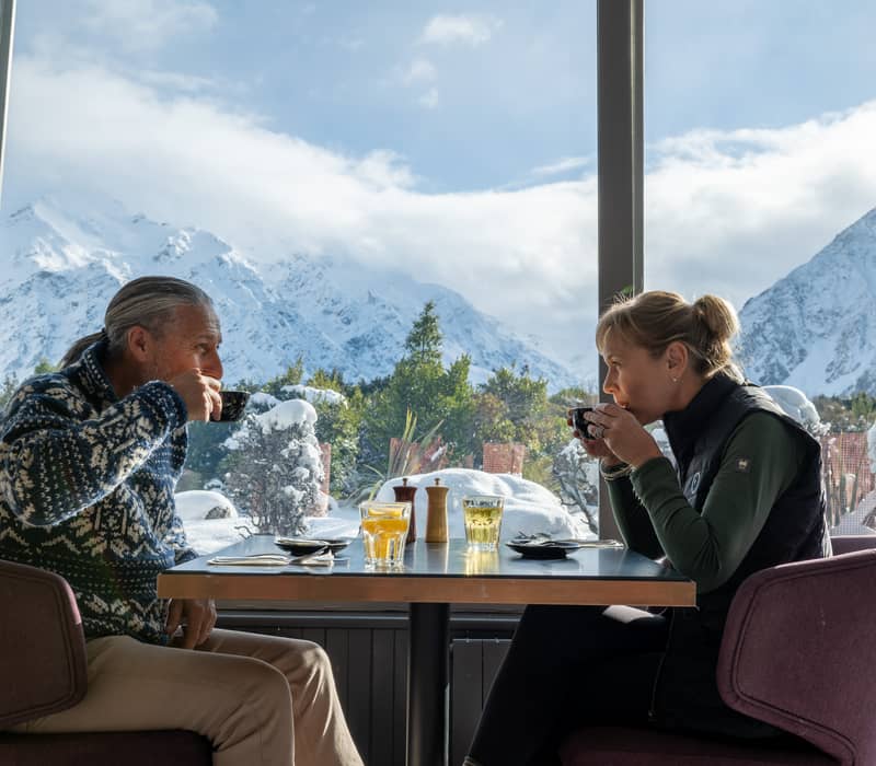 Couple drinking coffee inside a cafe with snow-covered mountains seen through the window.