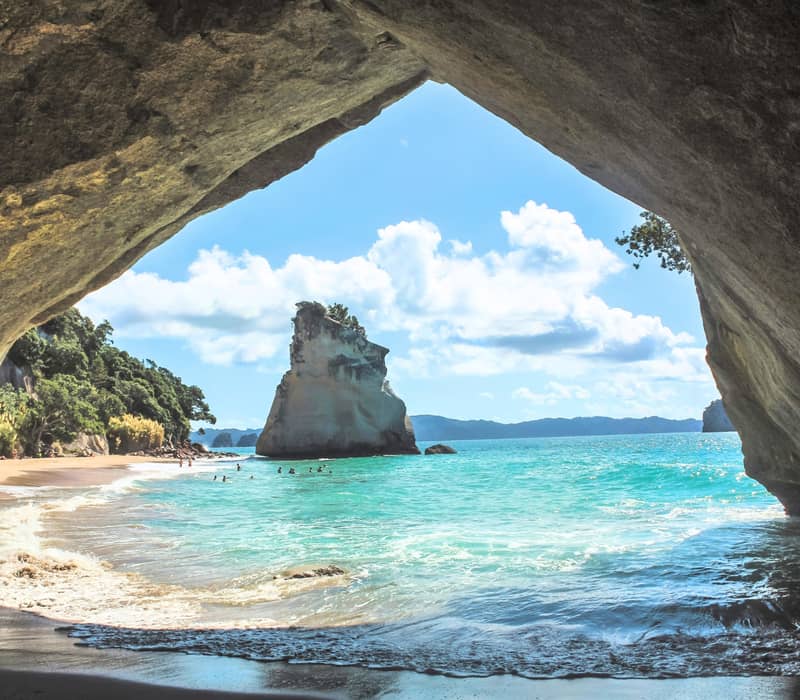 View from inside a cave of sandy beach, turquoise ocean, sea stack, and blue sky with clouds. Credit: Pure Discoveries