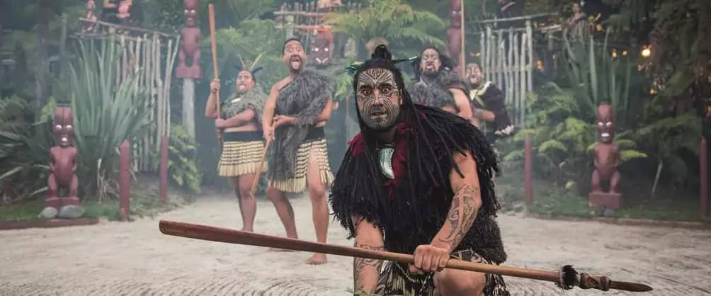 Maori warriors in traditional dress with tattoos perform a cultural dance in a forested outdoor area. Credit: Pure Discoveries