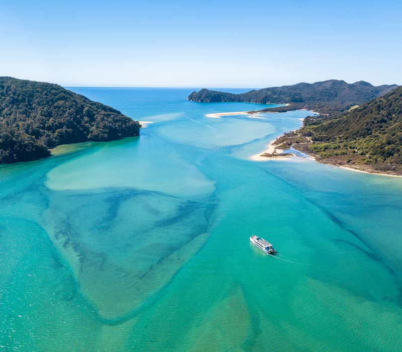 A tour boat sailing through clear turquoise water with forested coastline and sandy beaches. Credit: Pure Discoveries