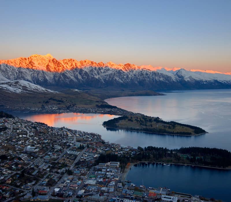 Sunset over Queenstown with pink snow-capped mountains, calm lake, and town buildings below. Credit: Pure Discoveries