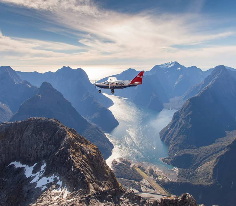 A small white plane with a red tail flying high above the jagged, snow-capped granite peaks of Milford Sound, with the sun reflecting off the water deep in the fiord below. Credit: Real Journeys