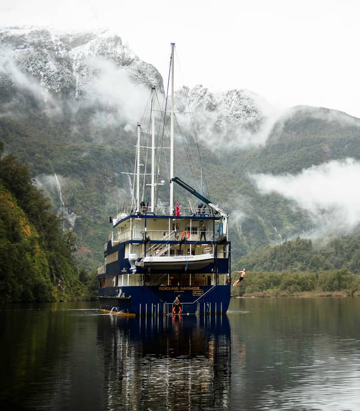 Large ship anchored in misty fjord with mountains and forests surrounding calm water. Credit: Real Journeys