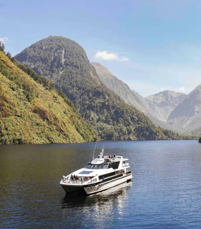 Boat sailing through calm fjord waters surrounded by green mountains under a clear sky. Credit: Real Journeys