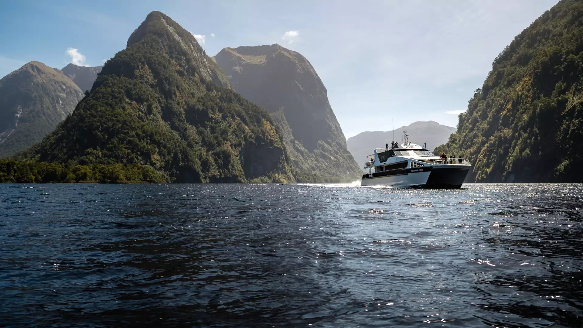 Boat sailing through calm fjord waters surrounded by green mountains under a clear sky. Credit: Real Journeys