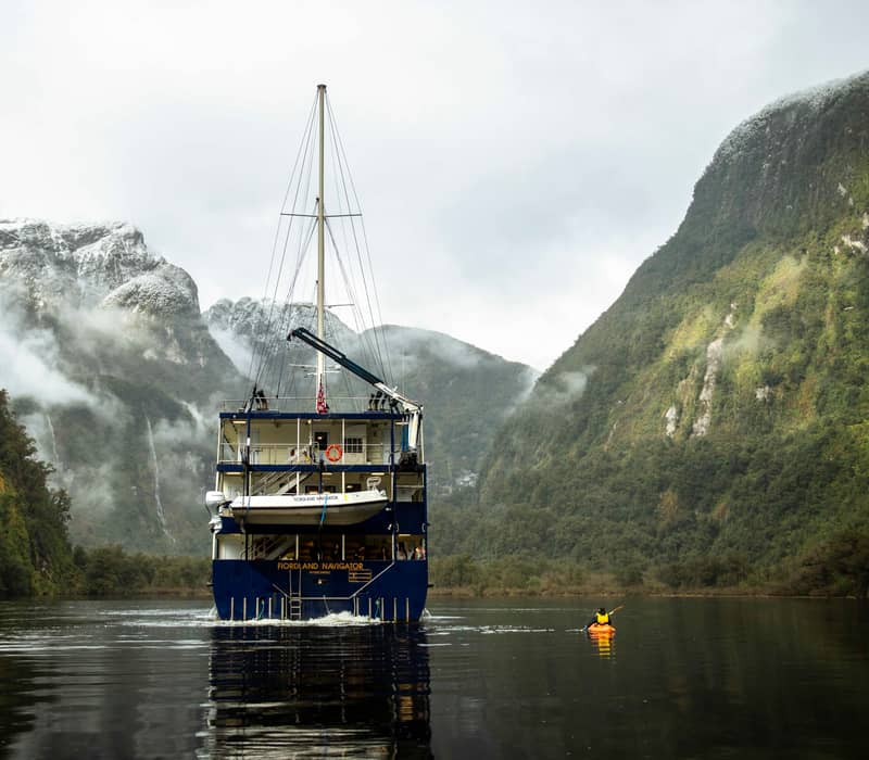 Cruise ship in fjord surrounded by foggy mountains and kayak on calm water Credit: Real Journeys