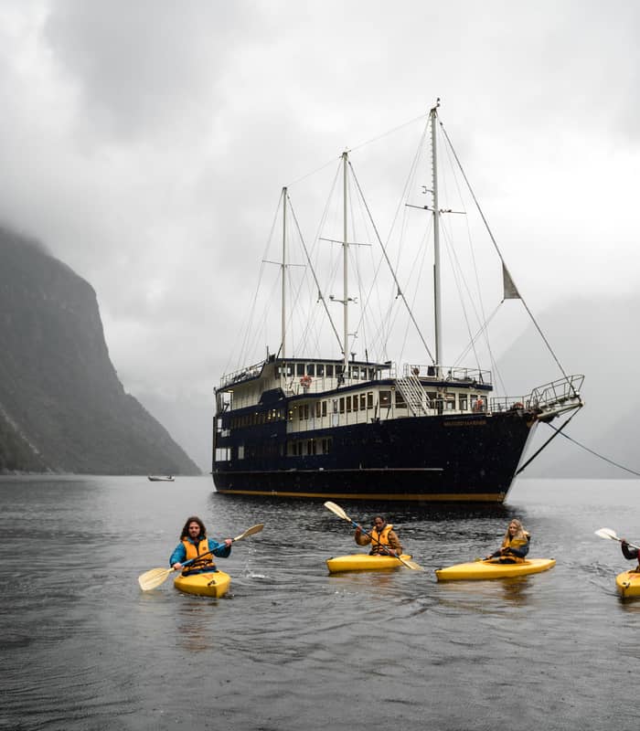 Four kayakers paddling in yellow kayaks near a cruise ship in a misty mountain fjord. Credit: Real Journeys