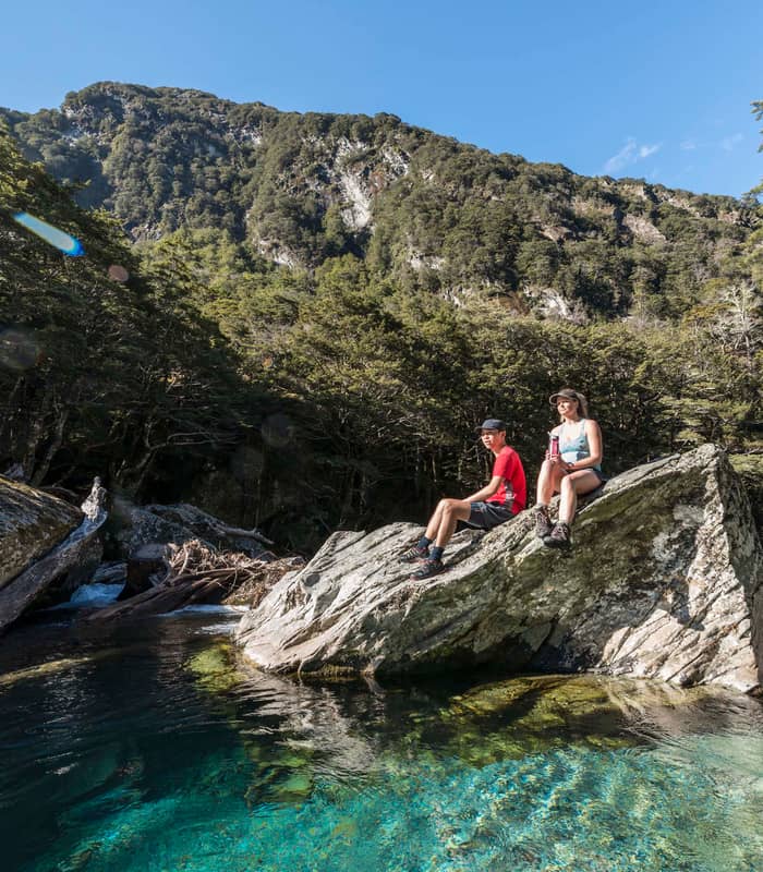 Two people sitting on rocks by a clear blue river with forest and mountains in background. Credit: Ultimate Hikes