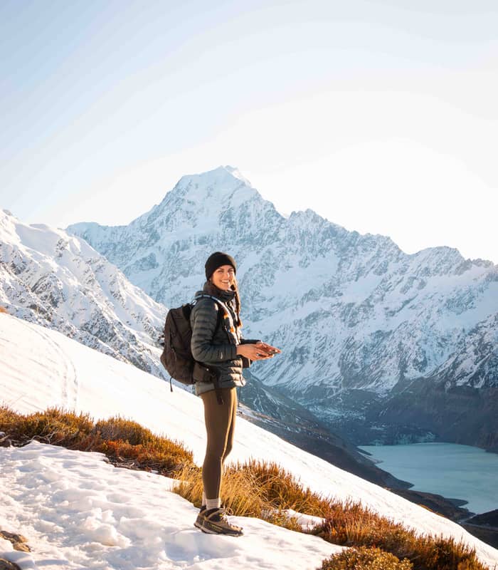 Woman standing on a snowy slope with backpack overlooking a lake and snow-covered mountains.