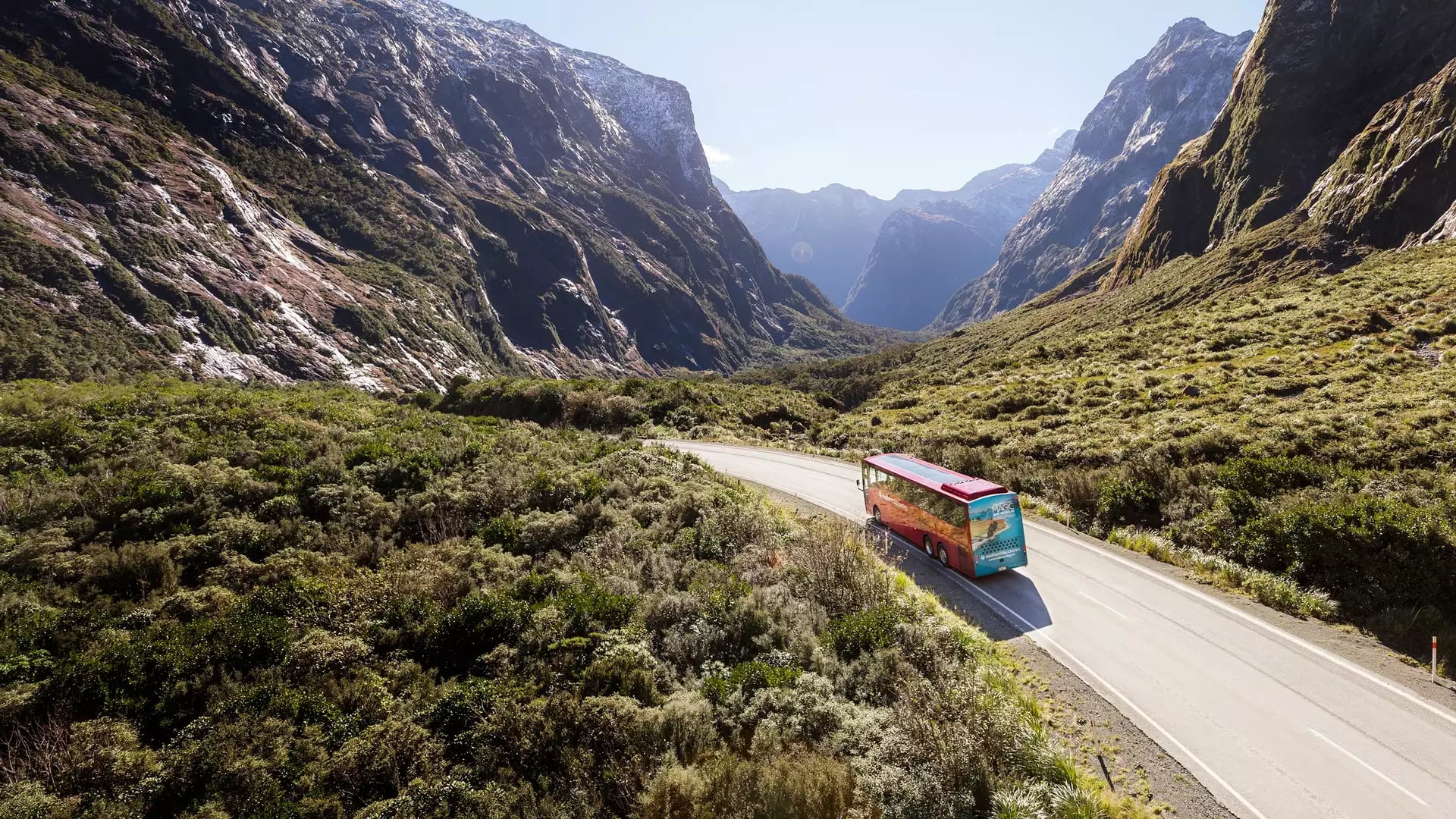 An elevated view of a red passenger coach driving along a winding road between steep forested mountain walls. Credit: Southern Discoveries