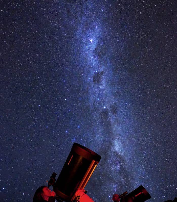 Two people looking through telescopes at the starry night sky with the Milky Way visible.