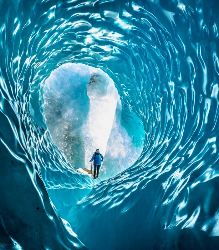 Person standing inside a blue ice cave with textured glacier walls.