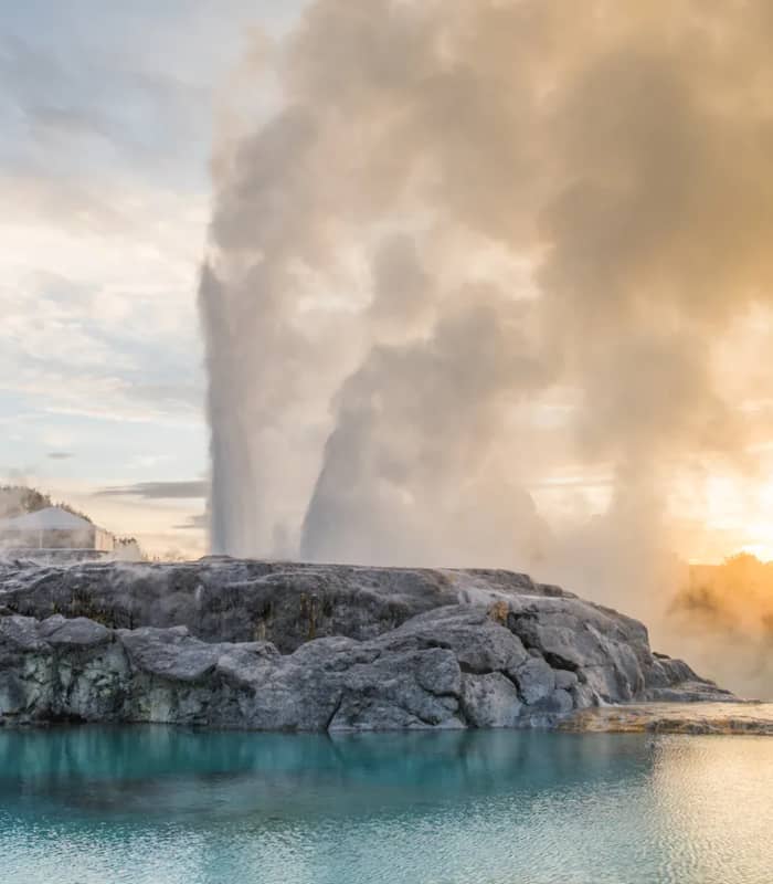 The Pōhutu Geyser erupts with a massive plume of steam and water behind a rocky silica terrace, reflected in a calm turquoise pool under a golden sky. Credit: Rotorua NZ - Media Library