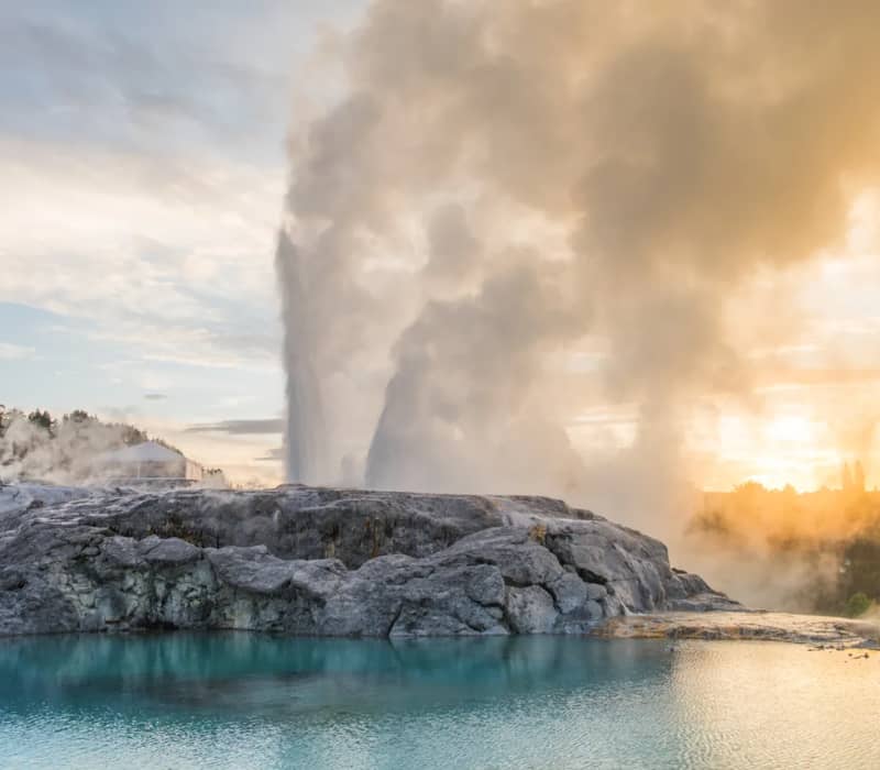 The Pōhutu Geyser erupts with a massive plume of steam and water behind a rocky silica terrace, reflected in a calm turquoise pool under a golden sky. Credit: Rotorua NZ - Media Library
