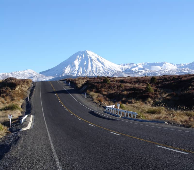 Paved road heading towards snow-covered Mount Ngauruhoe volcano under a clear blue sky. Credit: New Zealand Fine Touring
