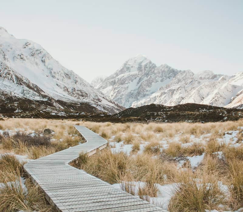 A wooden boardwalk winds through a snowy tussock valley leading toward the majestic snow-capped peak of Aoraki / Mount Cook. Credit: Jordan Heath - Unsplash