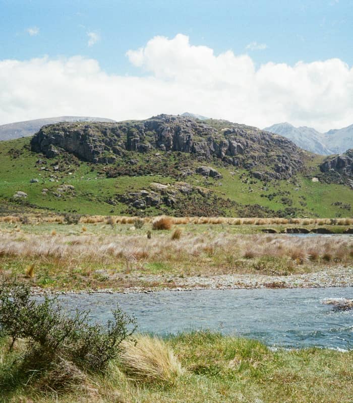 A wide landscape view of Mount Sunday, a distinctive rocky hill rising from a flat, golden tussock valley next to a winding alpine river, backed by rugged mountain ranges under a cloudy blue sky. Credit: Christopher Stites - Unsplash