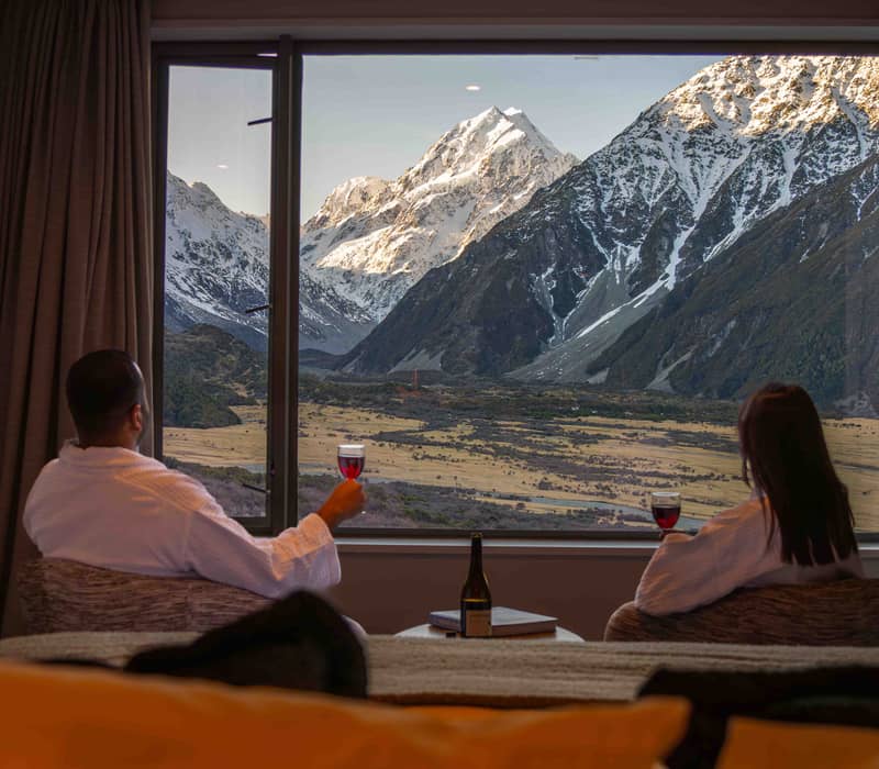 Couple in robes drinking wine looking at snowy mountain view through window