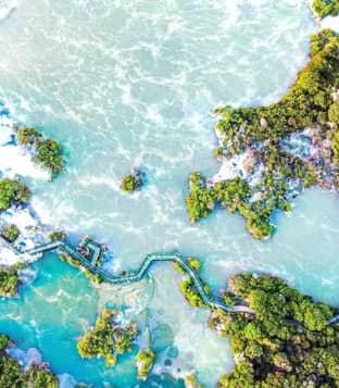 An aerial bird's-eye view of a winding wooden boardwalk crossing through vibrant turquoise and pale green geothermal pools surrounded by dense green native bush. Credit: Rotorua NZ - Media Library