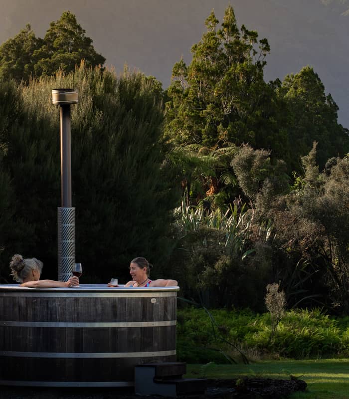 Two women in an outdoor wooden hot tub holding wine glasses with trees and hills behind them.