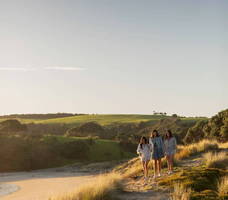 Three women walking along a sandy path on coastal dunes at sunset.