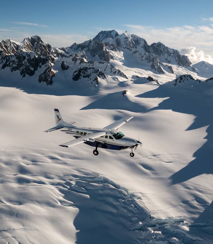 Small plane flying over snowy glacier and rugged mountain peaks under clear sky Credit: Air Safaris