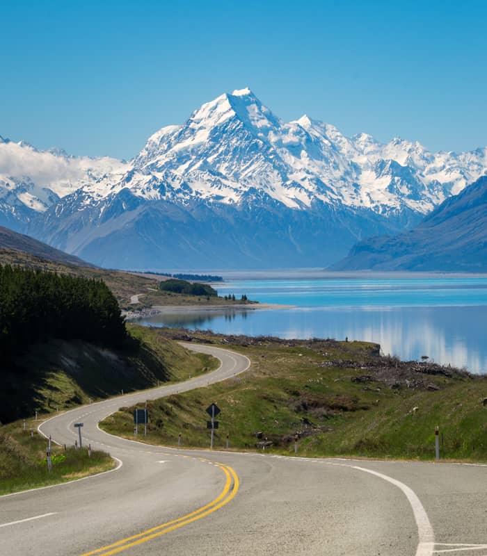 Curved road leading towards snow-covered Aoraki/Mt. Cook mountains by a reflective lake. Credit: New Zealand Fine Touring