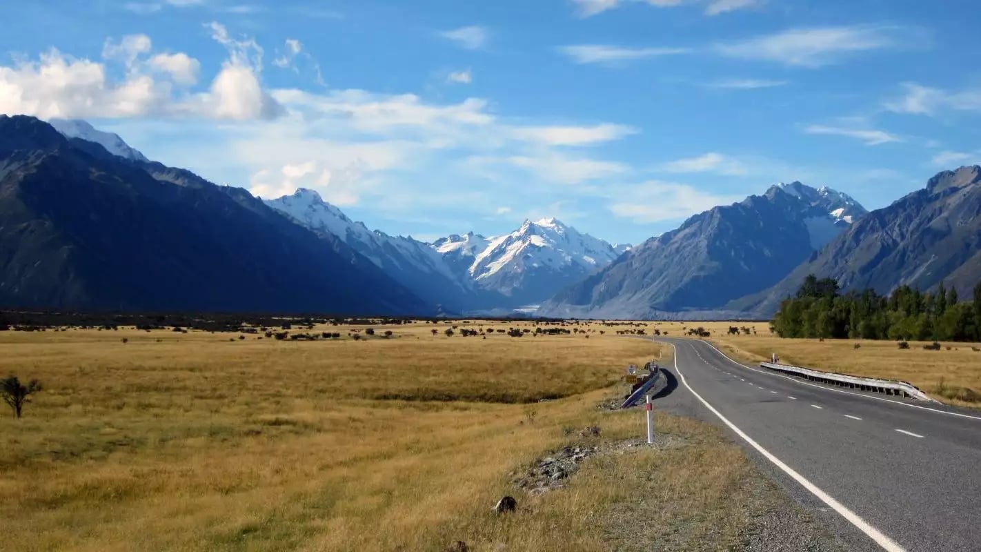 Road through grassland valley with snow-capped mountains under a blue sky with clouds. Credit: Cheeky Kiwi - Aoraki Mt Cook