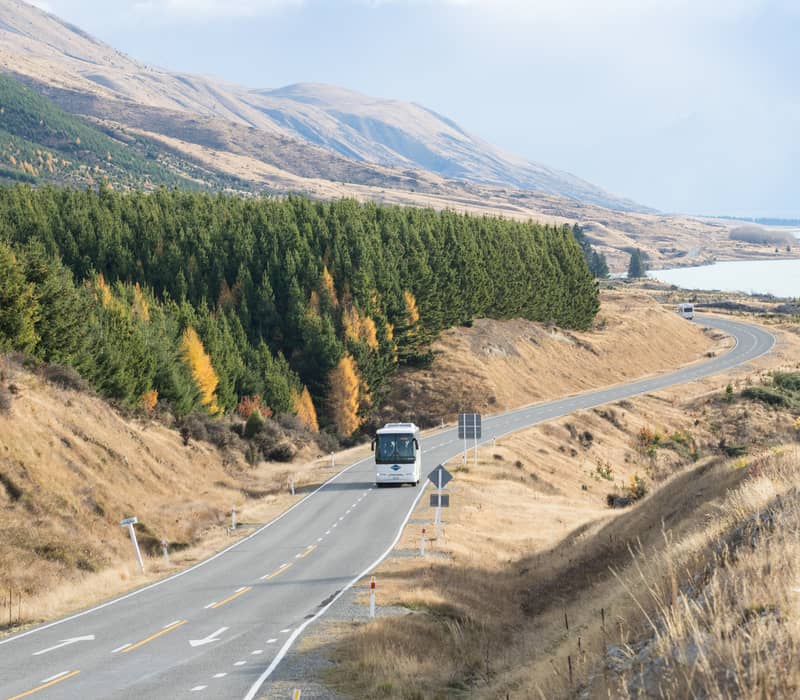 Coach driving on winding road next to Lake Pukaki with hills and trees showing autumn colors Credit: Entrada - Gray Line / Great Sights