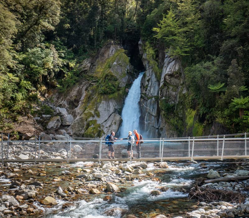 Three hikers standing on a swingbridge over a rocky river with a waterfall and forest in the background. Credit: Fiordland Trips & Tramps