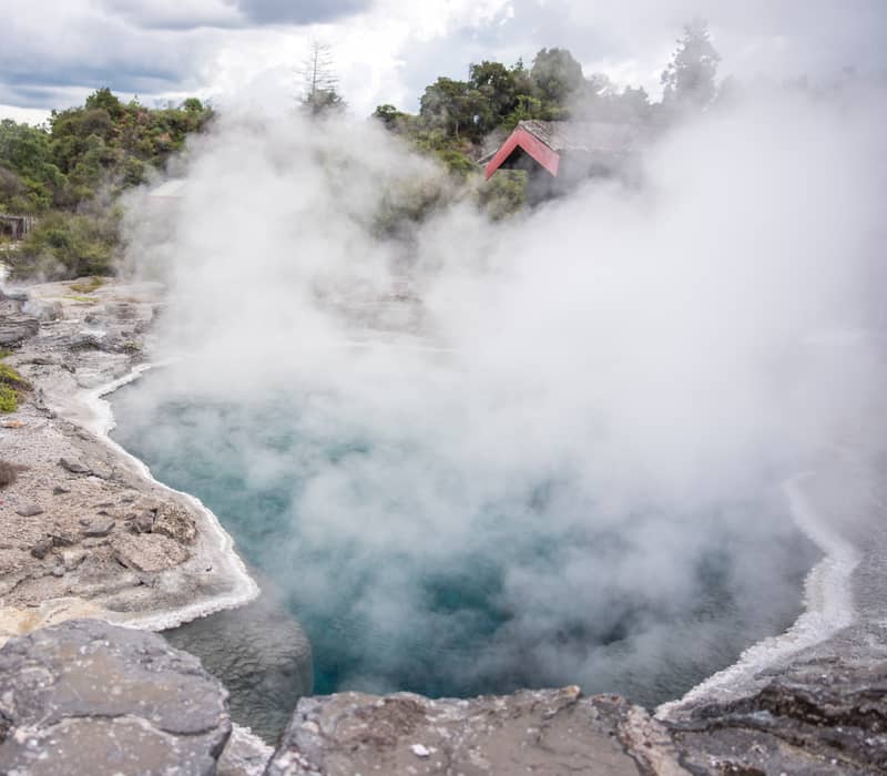 Steaming hot spring with rocky shoreline and surrounding greenery under cloudy sky. Credit: G Adventures