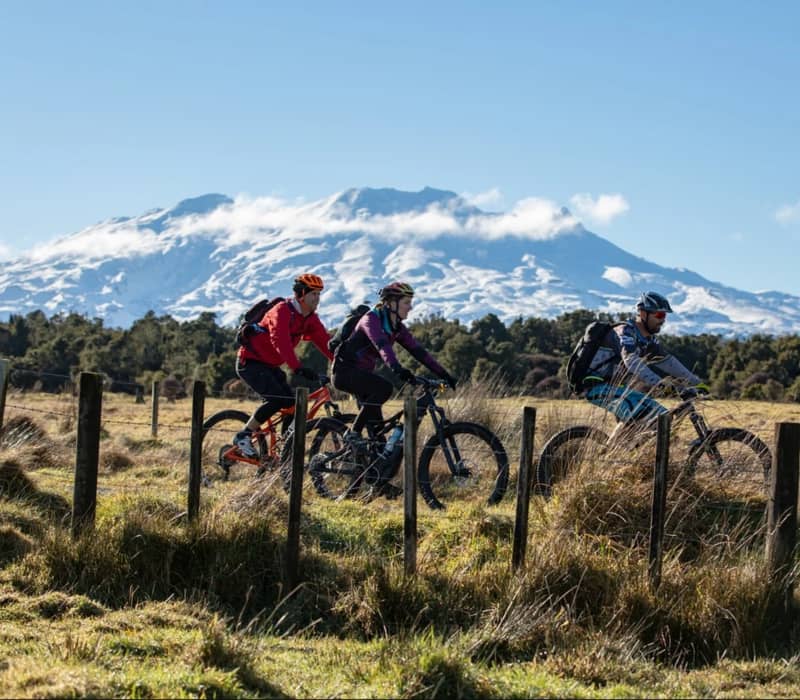 Three people biking with backpacks near a fence with snowy Tongariro mountain in the background. Credit: Great Journeys