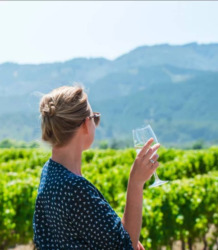 Woman holding a glass of white wine standing in a green vineyard with mountains in the background. Credit: Great Journeys