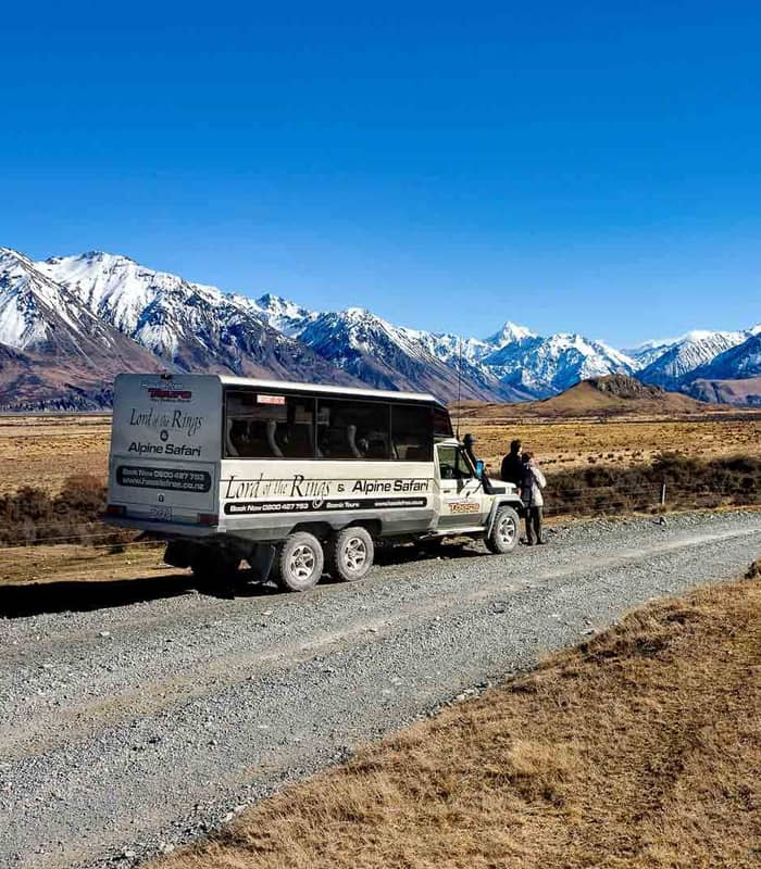 4x4 tour bus parked on a dirt road with people beside it and snow-covered mountains in the background under a clear sky. Credit: Hassle Free Tours