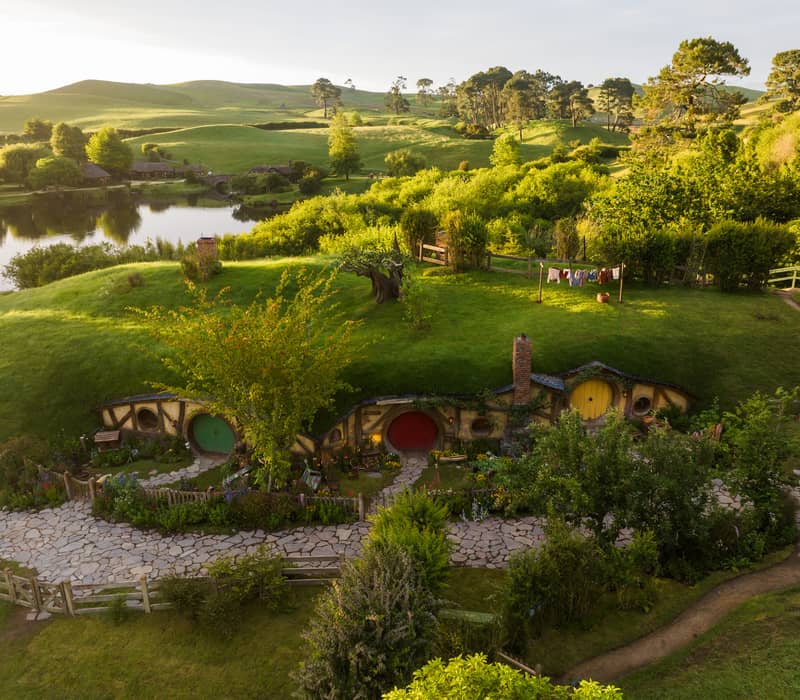 Hobbiton movie set with hobbit holes, gardens, and green hills beside a calm lake at sunset. Credit: Hobbiton Movie Set Tours
