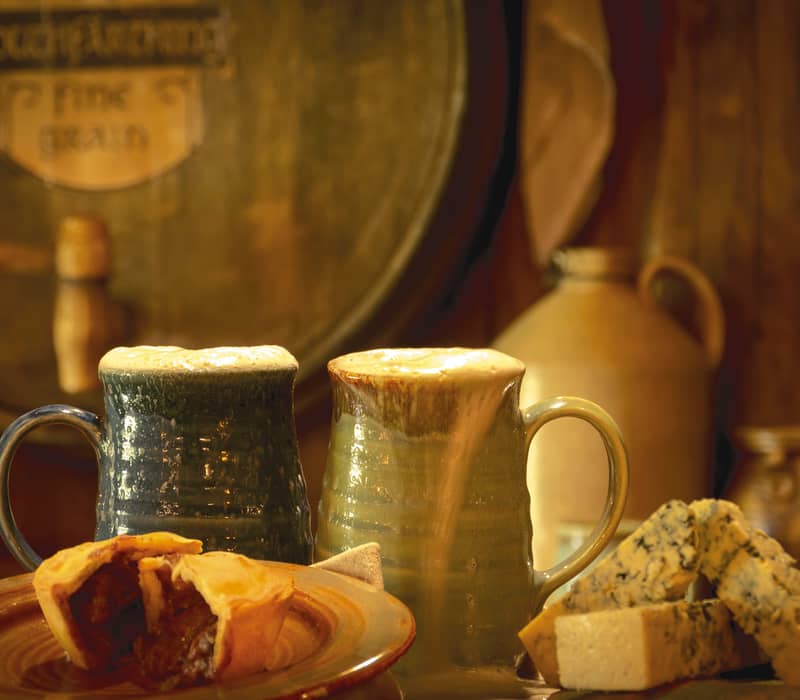 Two mugs of foamy beer with a meat pie and blue cheese on a wooden table. Credit: Cheeky Kiwi Travel