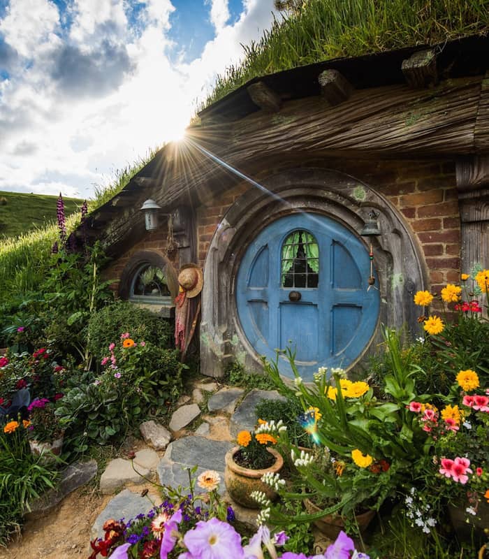 Hobbit house with blue round door and colorful garden under sunny sky with clouds. Credit: Hobbiton Movie Set Tours