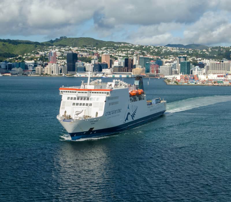 Ferry sailing in harbor with city and hills in the background under a cloudy sky. Credit: Interislander