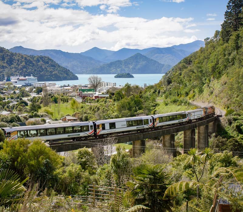 Passenger train crosses a viaduct with green hills and mountains in the background near water Credit: Kiwi Rail