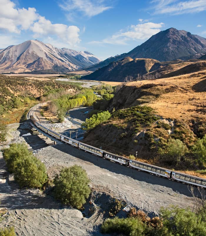 TranzAlpine train crossing a river bridge in a mountainous valley under blue sky and clouds Credit: Kiwi Rail