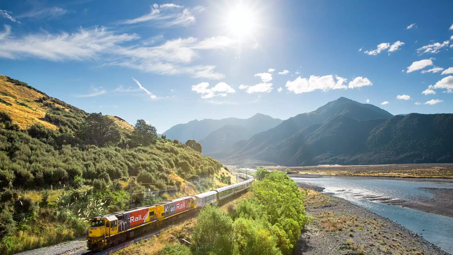 Scenic train crossing a bridge over the Waimakariri River with mountains in the background on a clear day. Credit: Kiwi Rail