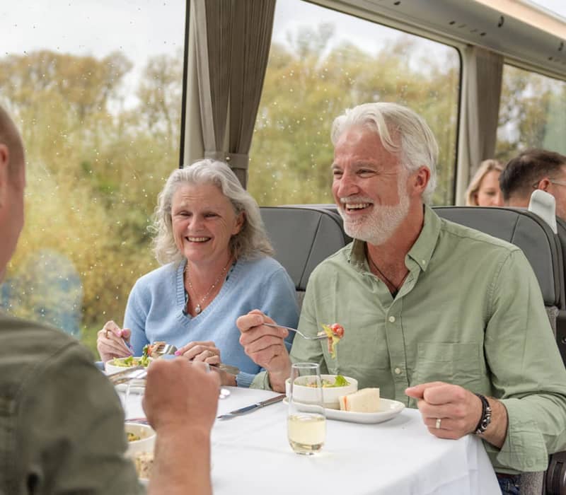A sunny daytime view of passengers smiling and laughing while enjoying a gourmet meal and regional wine inside the premium Scenic Plus carriage on the KiwiRail TranzAlpine train. Credit: Kiwi Rail