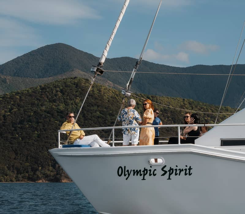 People on a sailboat named Olympic Spirit with mountains and water in the background on a sunny day. Credit: Marlborough Tour Company