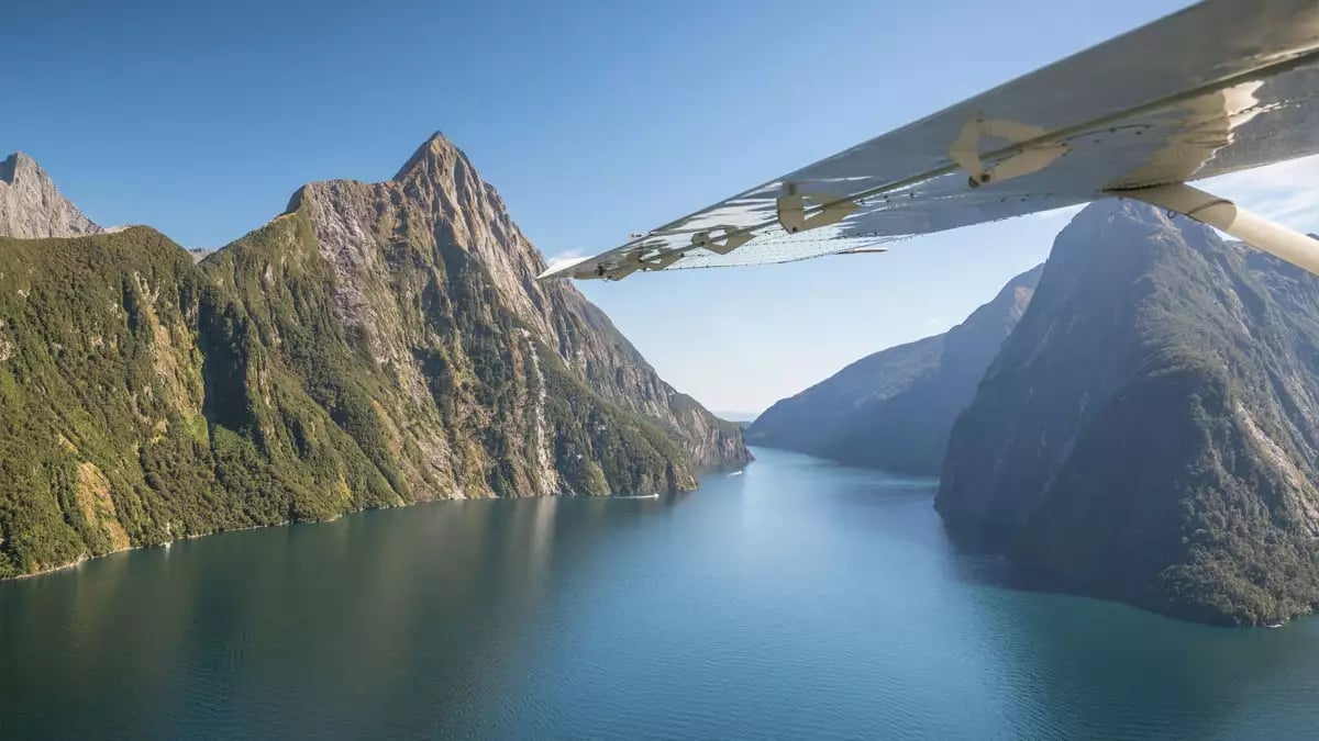 Aerial view of Milford Sound with mountains, water, cruise boats, and airplane wing under clear sky. Credit: Cheeky Kiwi Travel