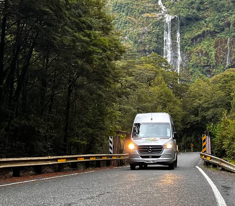Van on road through forest with waterfall and misty mountain behind Credit: Cheeky Kiwi Travel