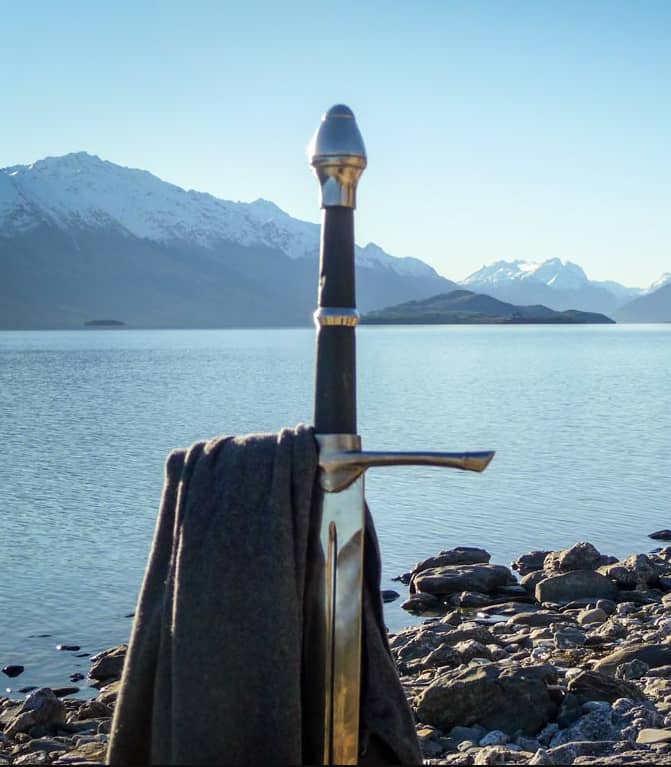 Sword with scarf on rocky shore by lake with snow-capped mountains. Credit: Nomad Adventures
