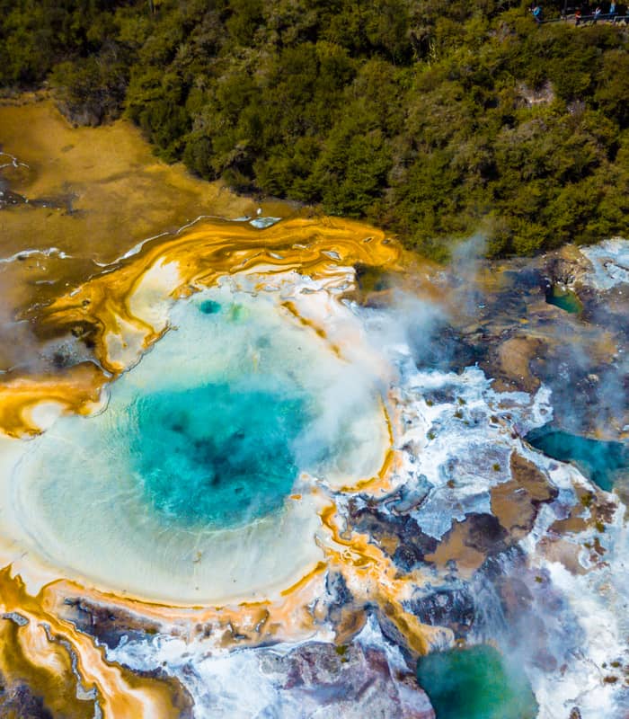Aerial view of a colorful geothermal hot spring with steam and forest around it.