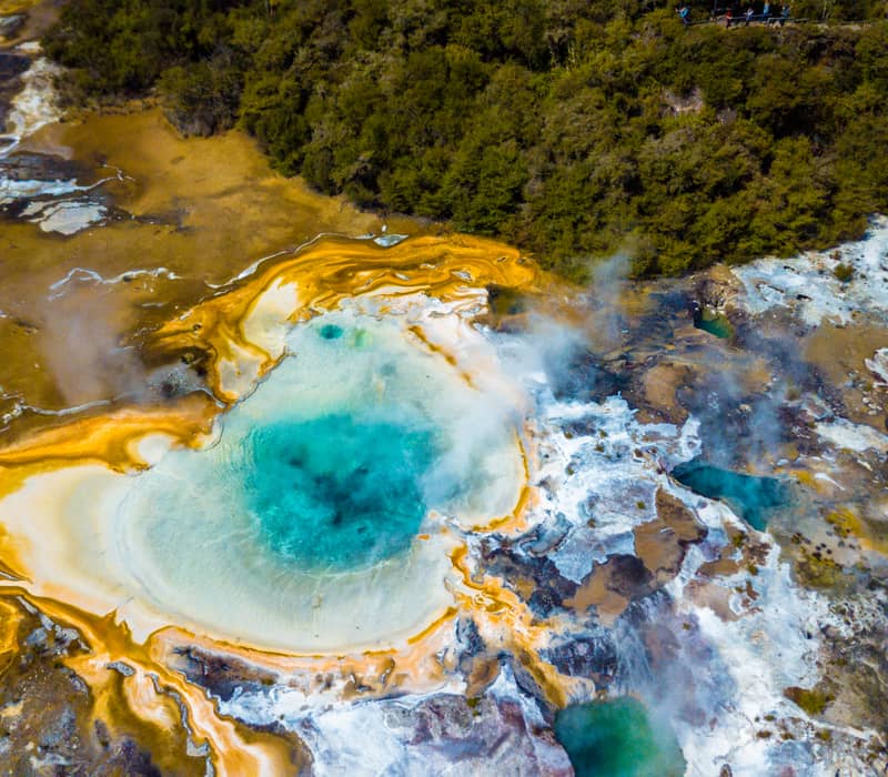 Aerial view of a colorful geothermal hot spring with orange and blue mineral deposits emitting steam near forest. Credit: New Zealand Fine Touring