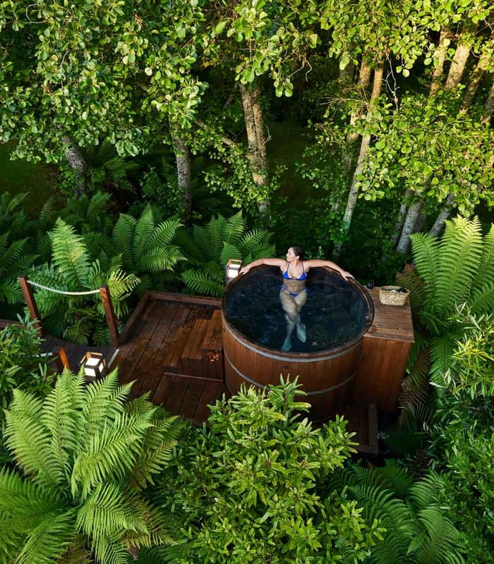 Woman in blue swimsuit relaxing in wooden hot tub surrounded by lush green ferns and trees on a deck. Credit: Secret Spot Hot Tubs Rotorua