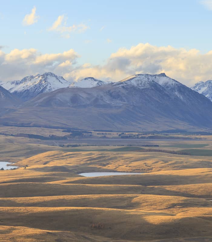 Rolling golden tussock hills with scattered small lakes, backed by a grand, snow-capped mountain range under a soft, cloudy sky. Credit: Narongsak Nagadhana - Shutterstock